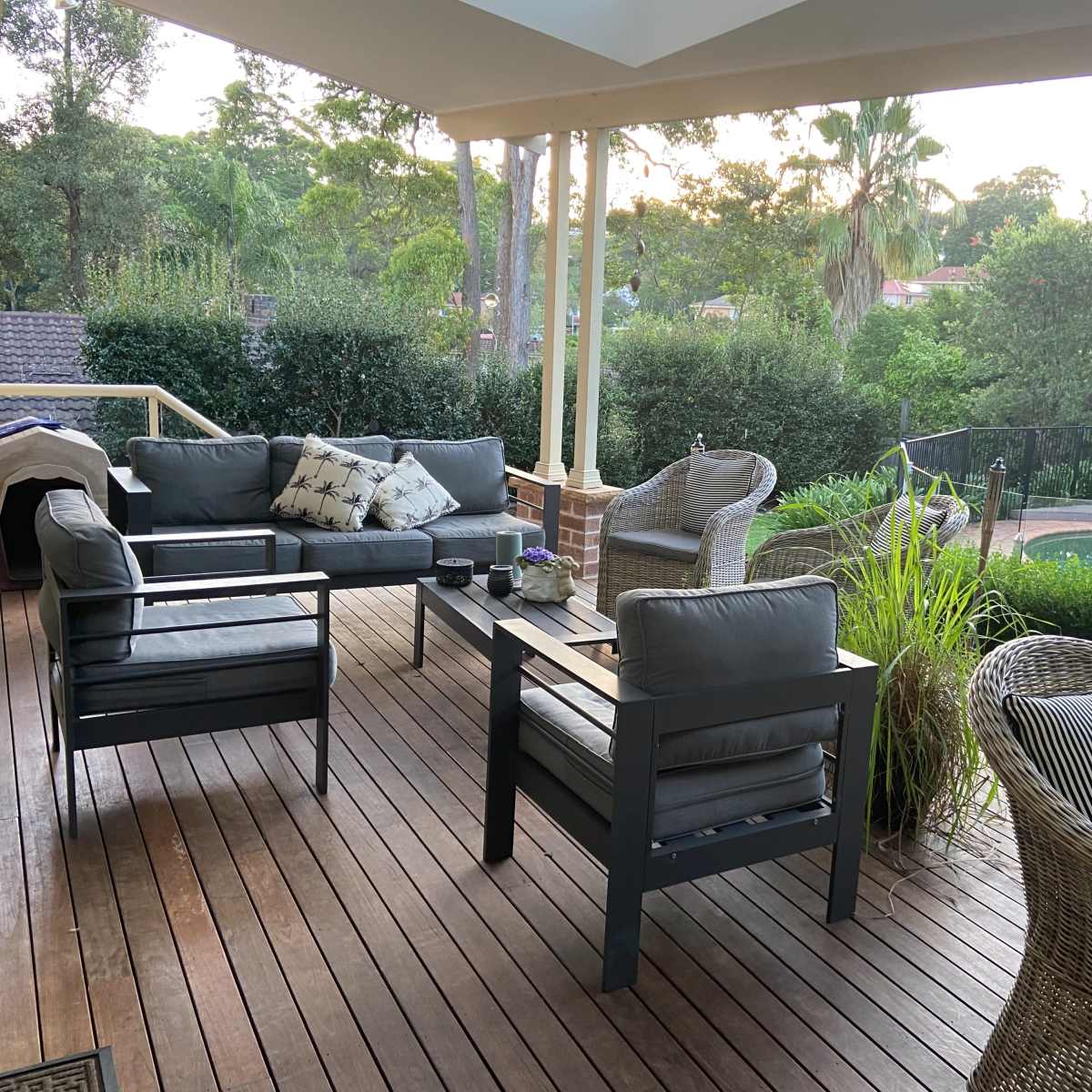 A patio features a seating area with gray couches and a wicker chair, surrounded by greenery and overlooking a swimming pool.