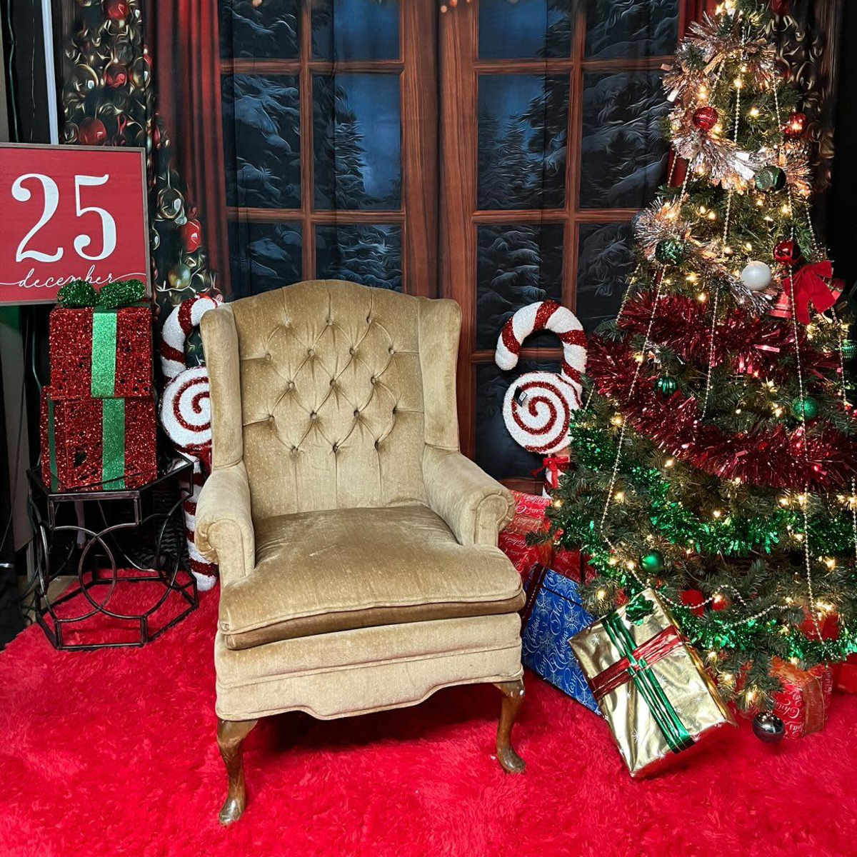 A decorated Christmas scene with a green and red festive tree, presents, a large red sign displaying "25," and a beige tufted armchair set against a backdrop of snow-covered windows.