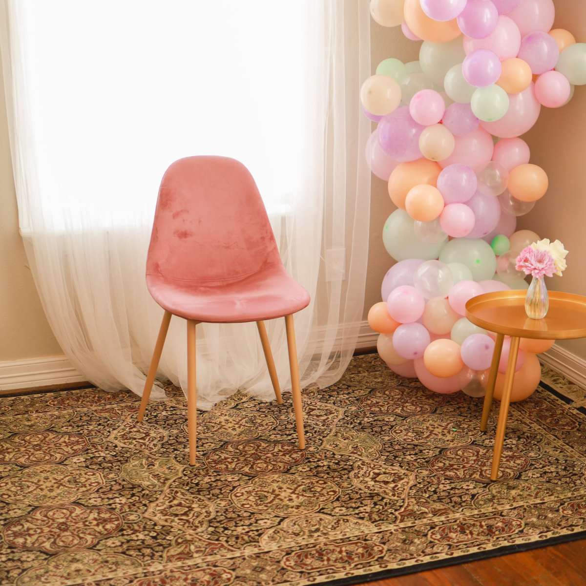 A pink chair stands next to a colorful balloon arrangement in a softly lit room with a window draped in sheer curtains.