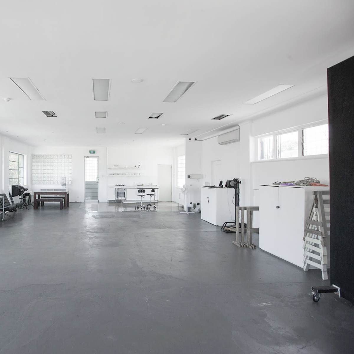 A spacious, minimalist studio with white walls, large windows, and various furniture items scattered throughout the floor.