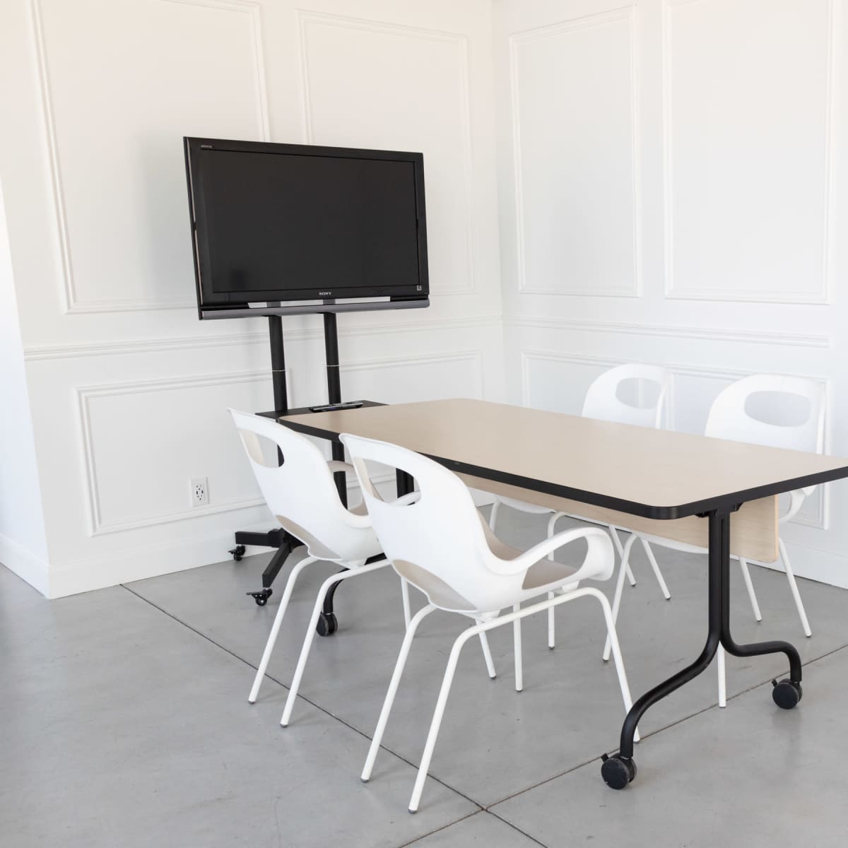 A conference room features a rectangular table surrounded by four white chairs and a mounted television on a movable stand.