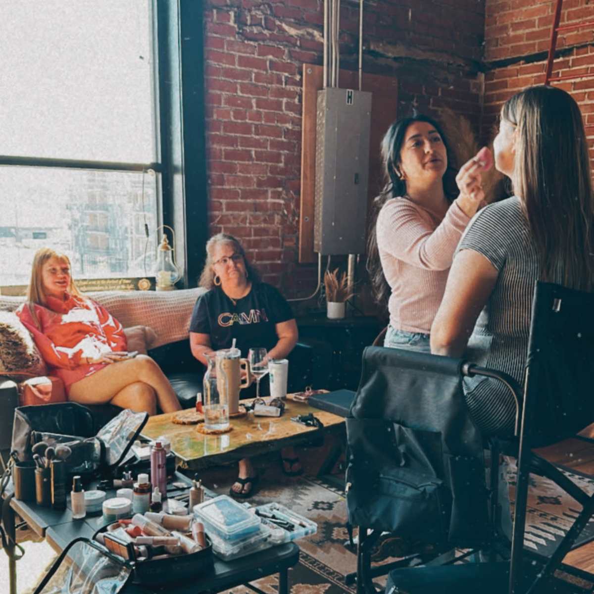 A group of four women is gathered in a room with exposed brick walls, where one woman is applying makeup to another while two others sit nearby on a couch.