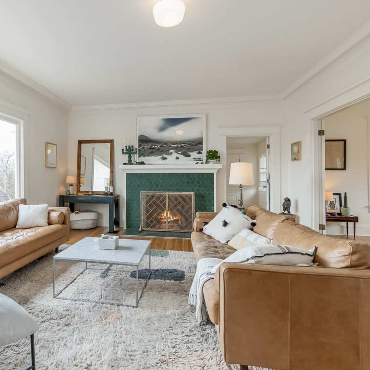A spacious living room features two leather sofas, a coffee table, a fireplace with a green-tiled surround, and large windows allowing natural light to enter.
