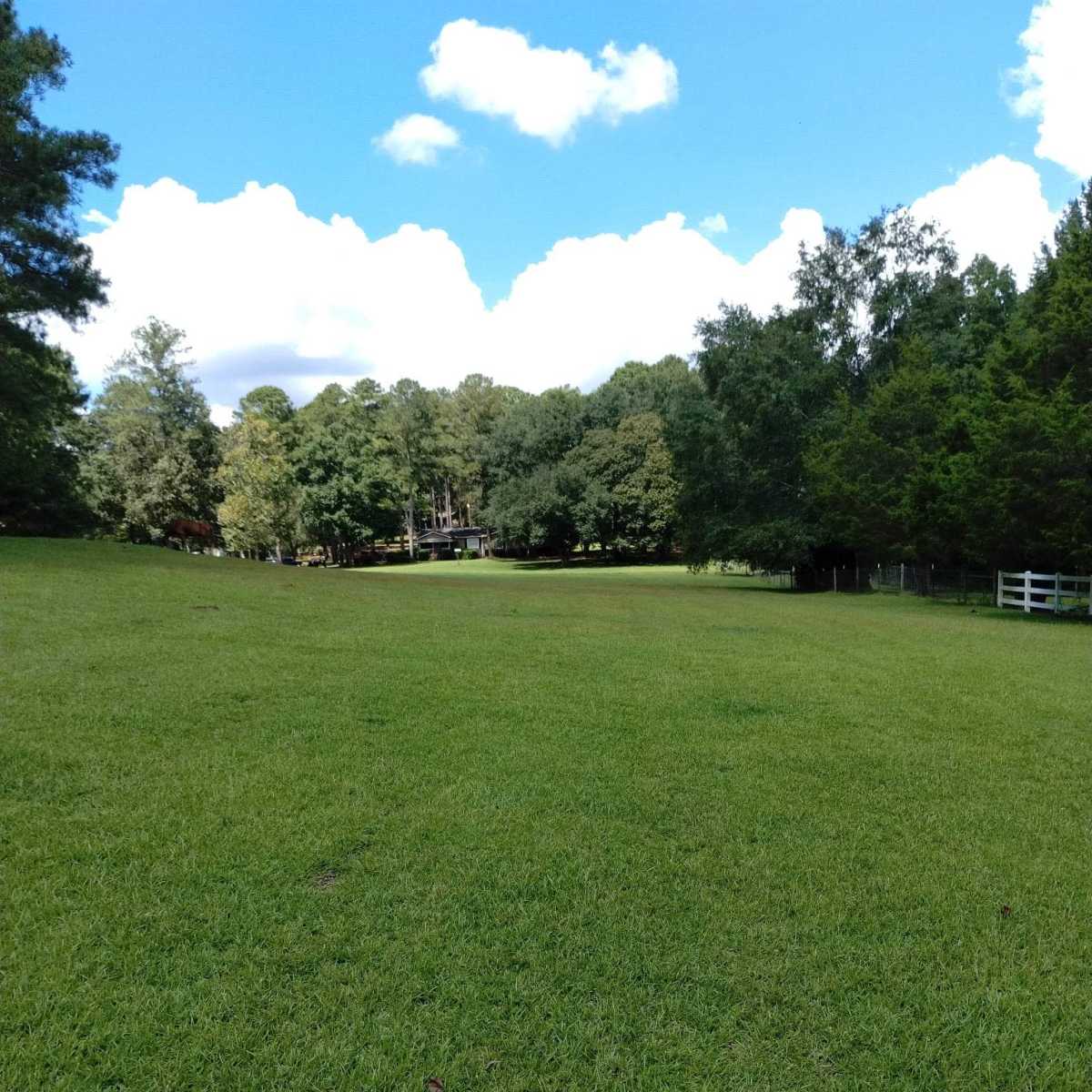 A wide green field extends towards a house nestled among trees under a partly cloudy sky.