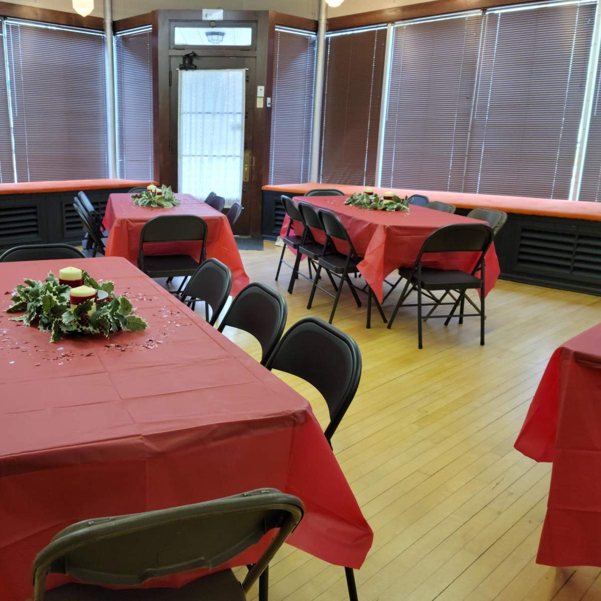 The room features several tables covered with red tablecloths, arranged with centerpieces of greenery, and is illuminated by natural light from large windows.