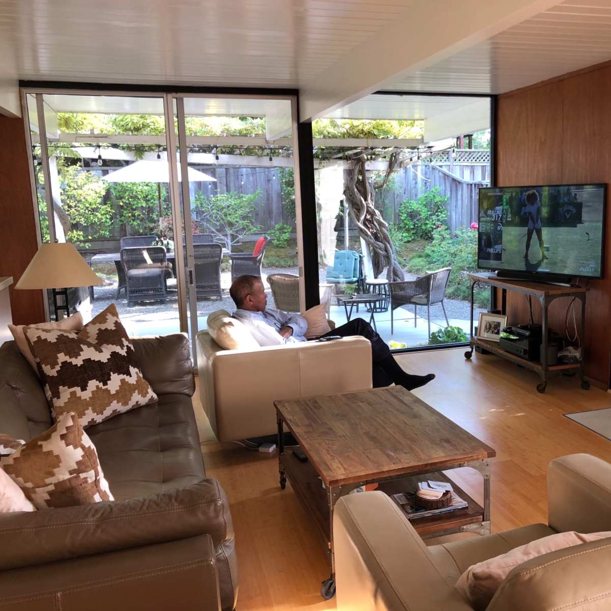 A man sits on a couch watching television in a modern living room with large windows overlooking a patio.