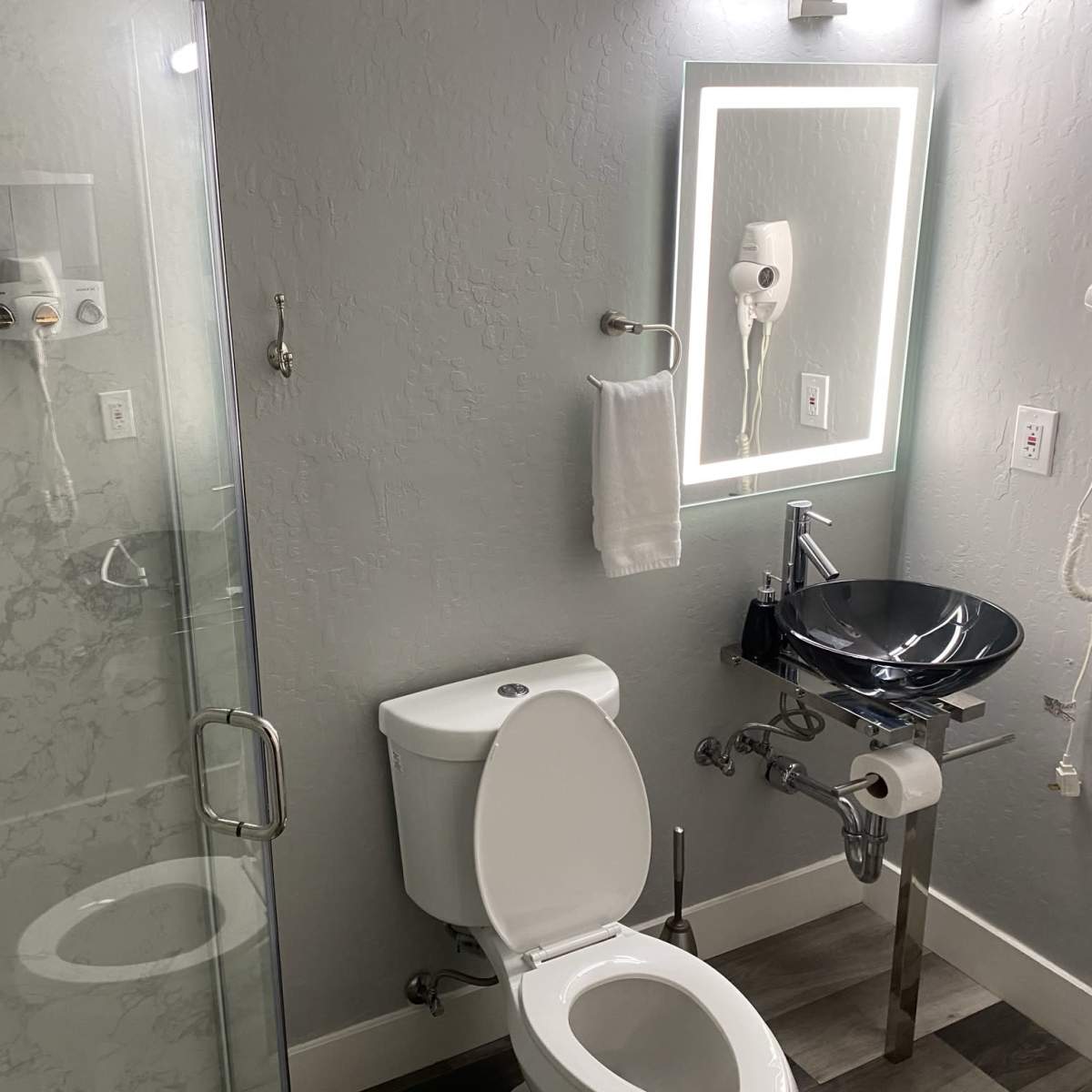 A modern bathroom featuring a glass shower, a white toilet, a sleek black sink, and a backlit mirror on a light gray wall.