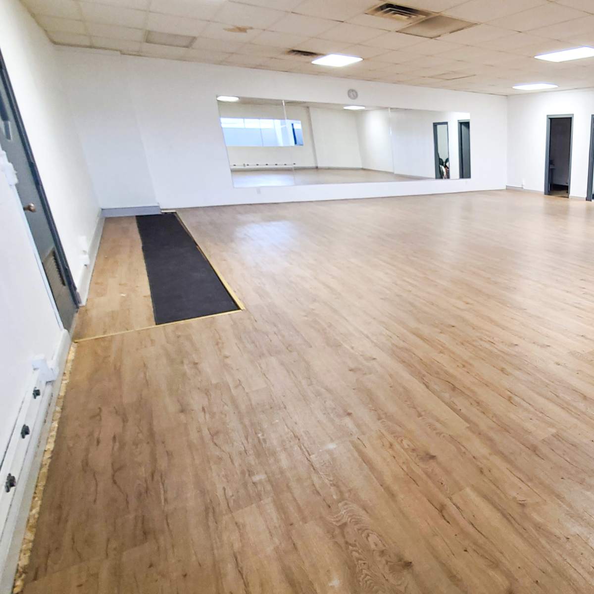 A large, empty room with wooden flooring, plain white walls, and mirrors on one side.