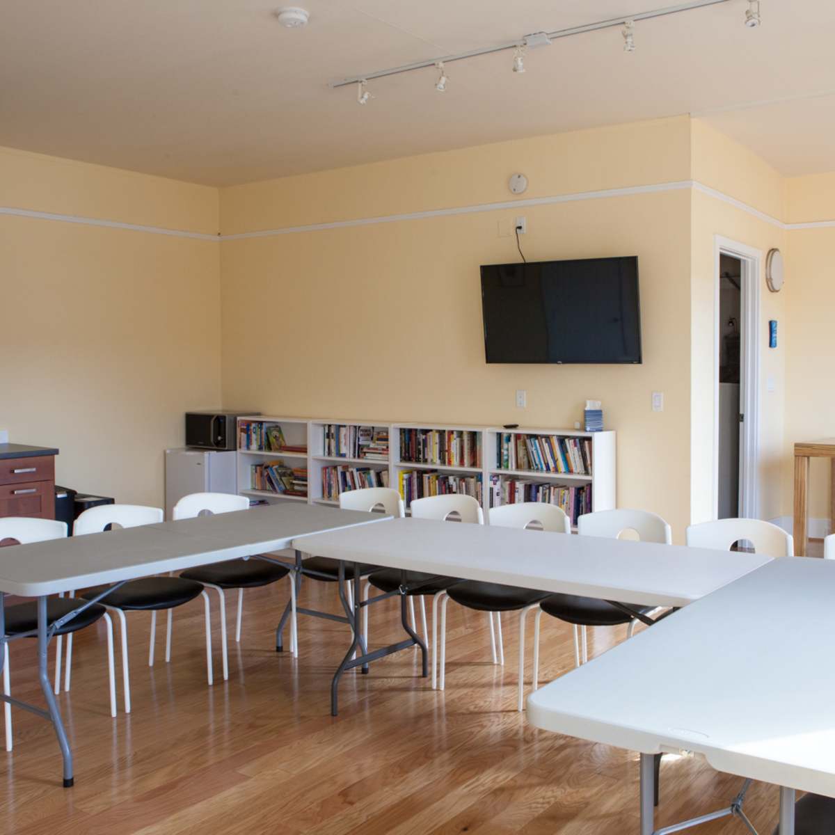 A multi-purpose room features a set of tables arranged in a U-shape with chairs, a kitchenette, a wall-mounted television, and a bookshelf.