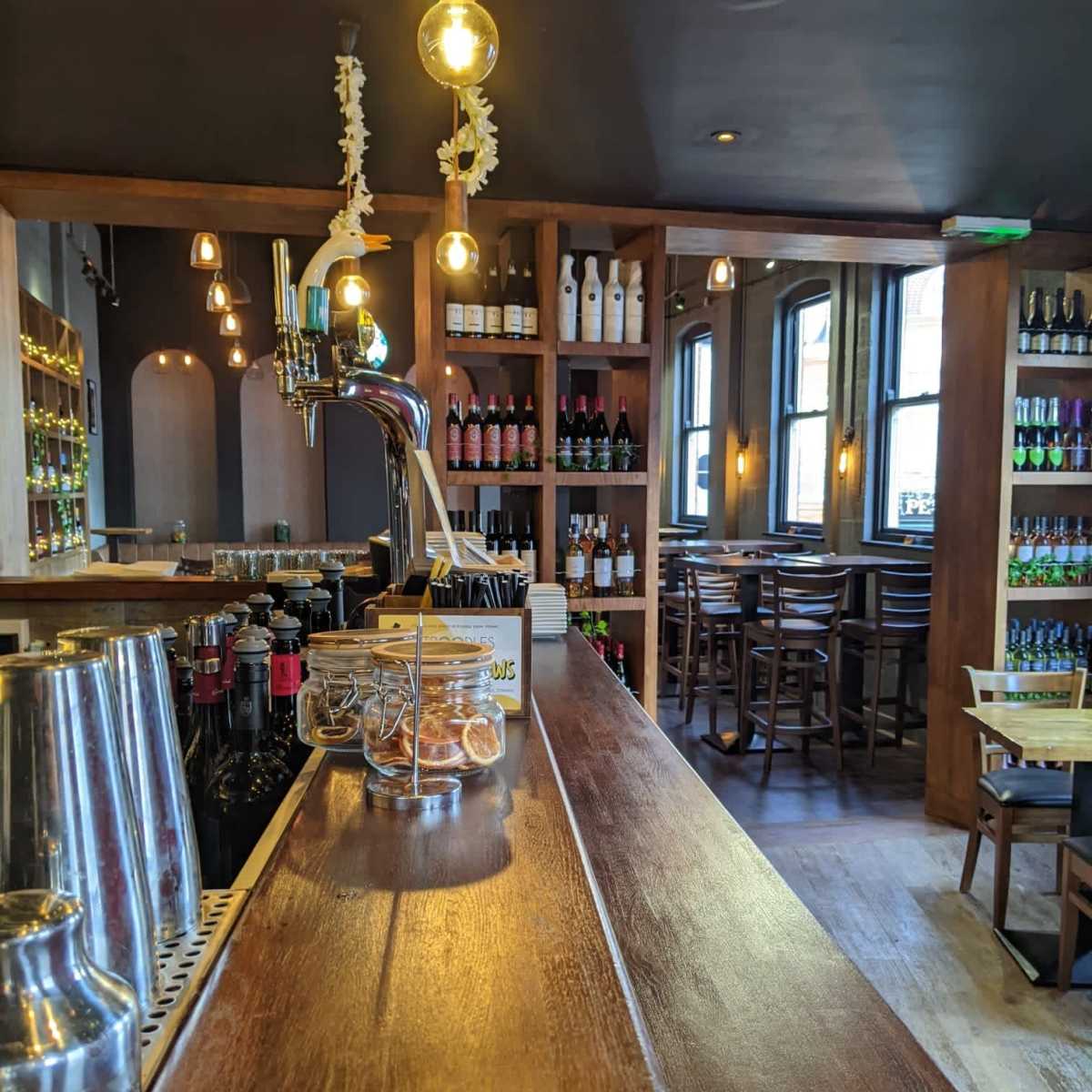 A wooden bar with a beer tap and shelves filled with bottles of wine and beer, set in a cozy interior with bar stools and warm lighting.