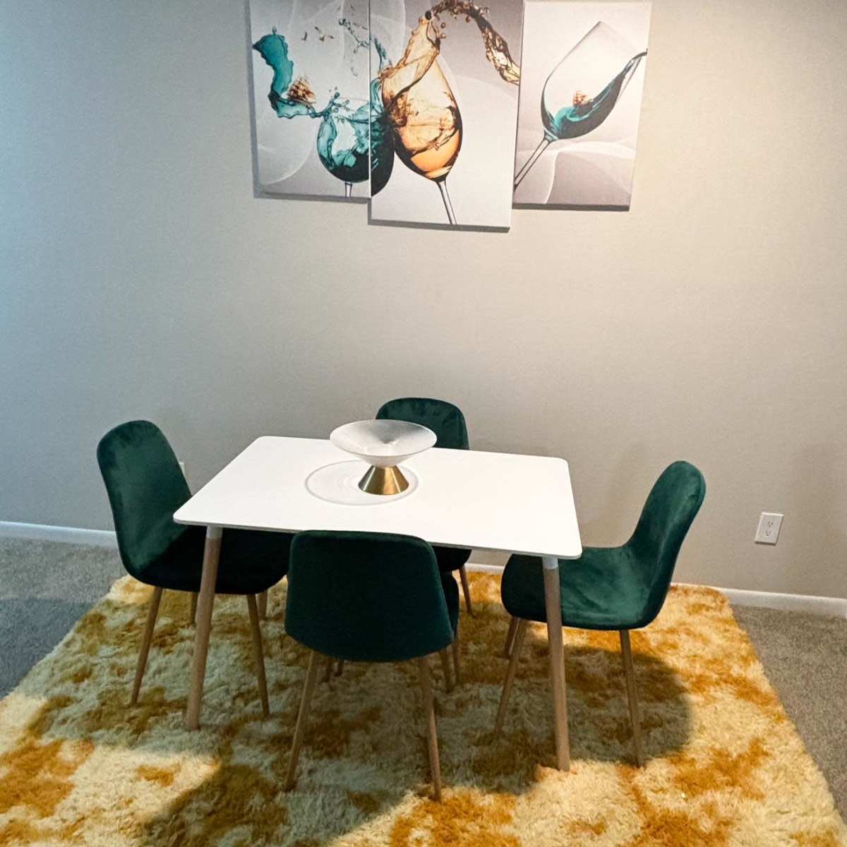 A modern dining area features a square white table surrounded by four green chairs on a large, yellow and cream rug, with abstract art depicting glasses of beverages on the wall.