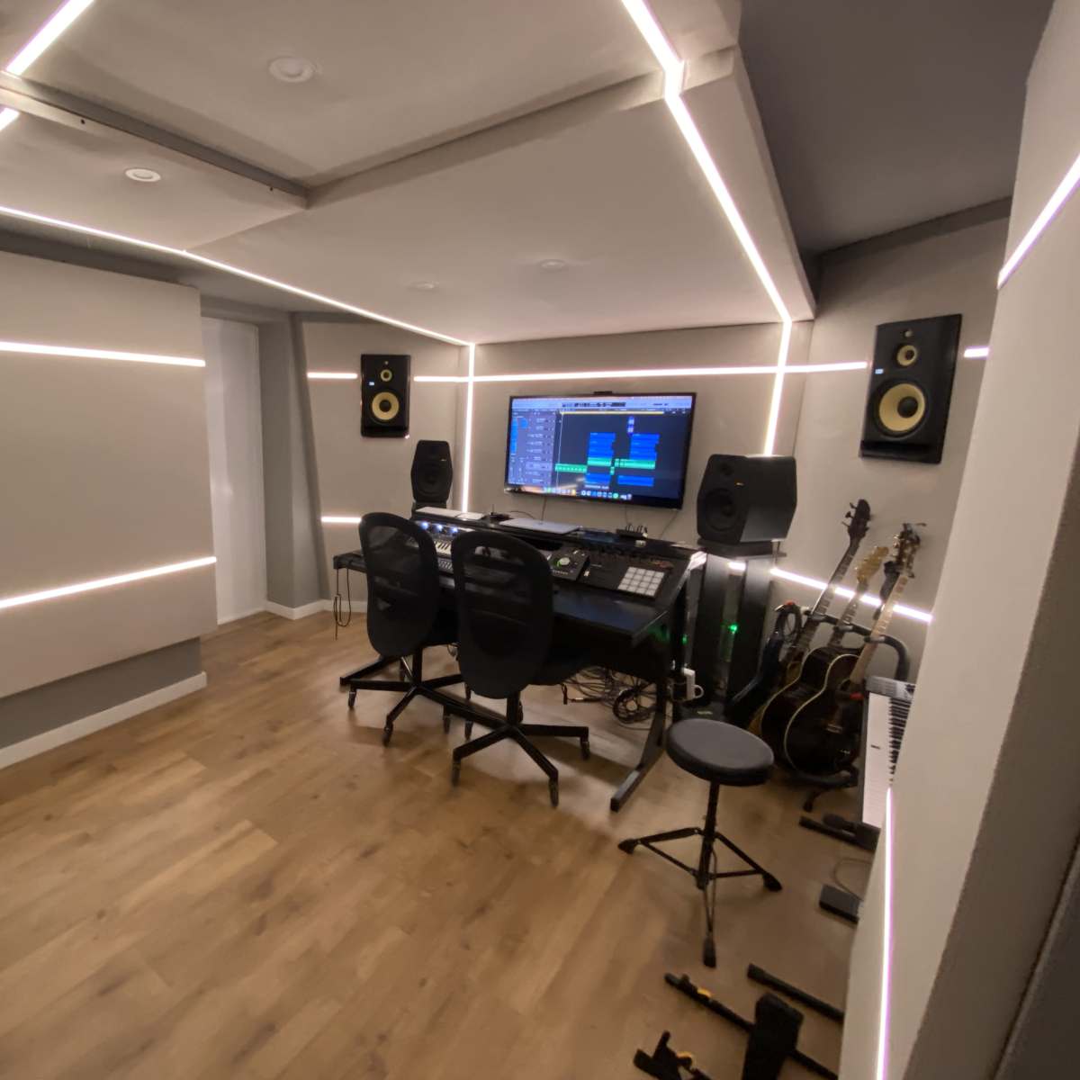 A music studio with a digital audio workstation setup, speakers, a seating area, and several guitars leaning against the wall.