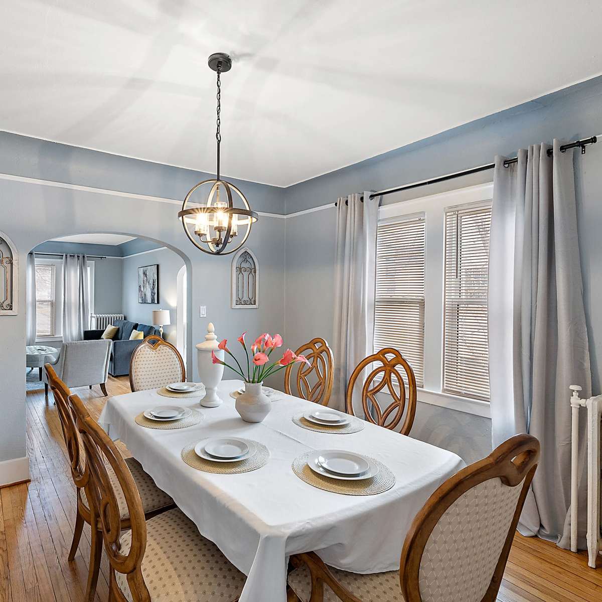 A dining area features a table set for six, surrounded by wooden chairs, with a chandelier overhead and a view into a living room through an arched entry.