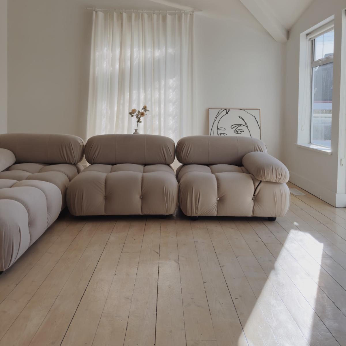 A light-filled living space features a beige, bulbous sofa, wooden floorboards, and sheer curtains beside a framed artwork.