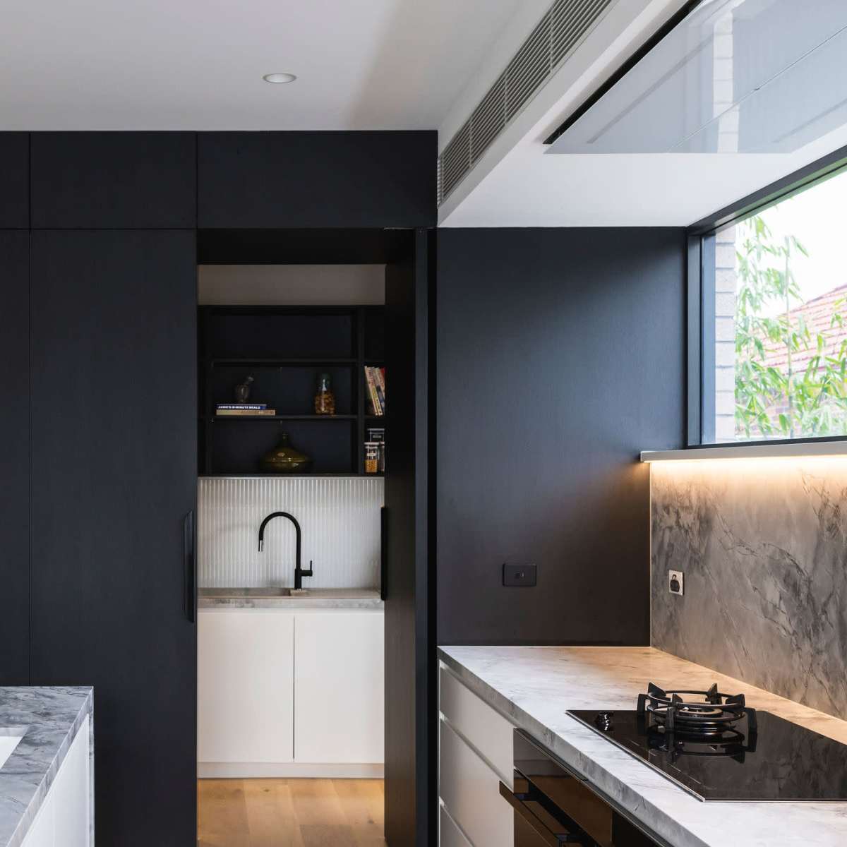 A modern kitchen with sleek black cabinetry, a marble countertop, and a small, open pantry area visible in the background.