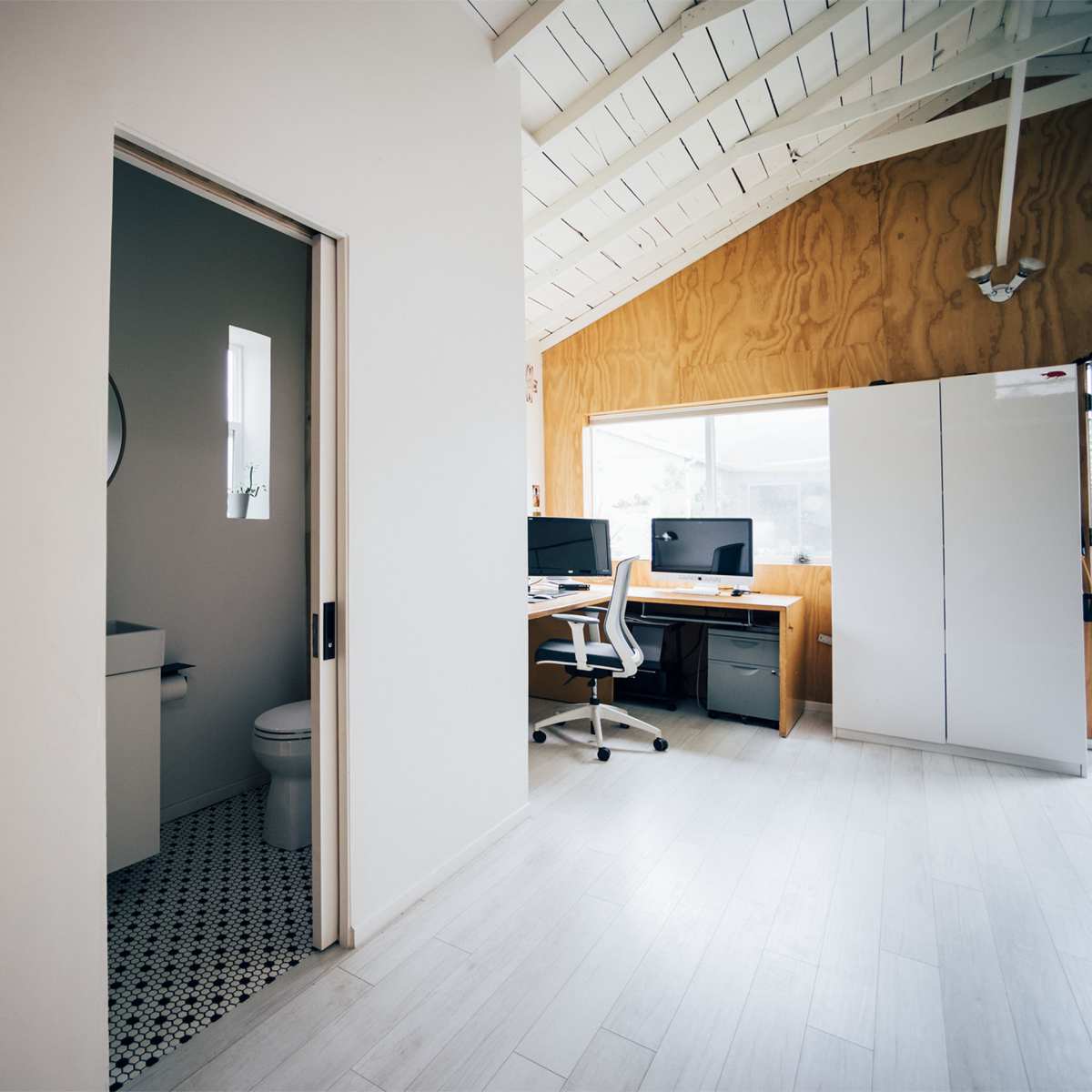 A modern interior space with a bathroom doorway on the left and a workspace featuring two computer desks on the right.