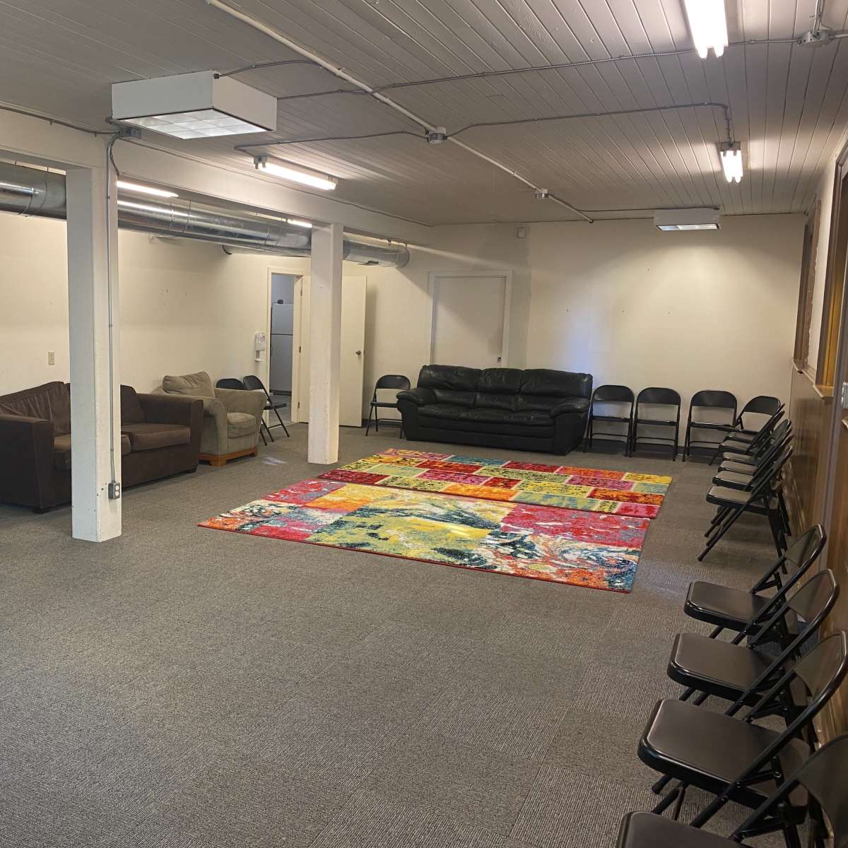 The room features a colorful area rug in the center, surrounded by rows of black folding chairs and a few couches along the walls.