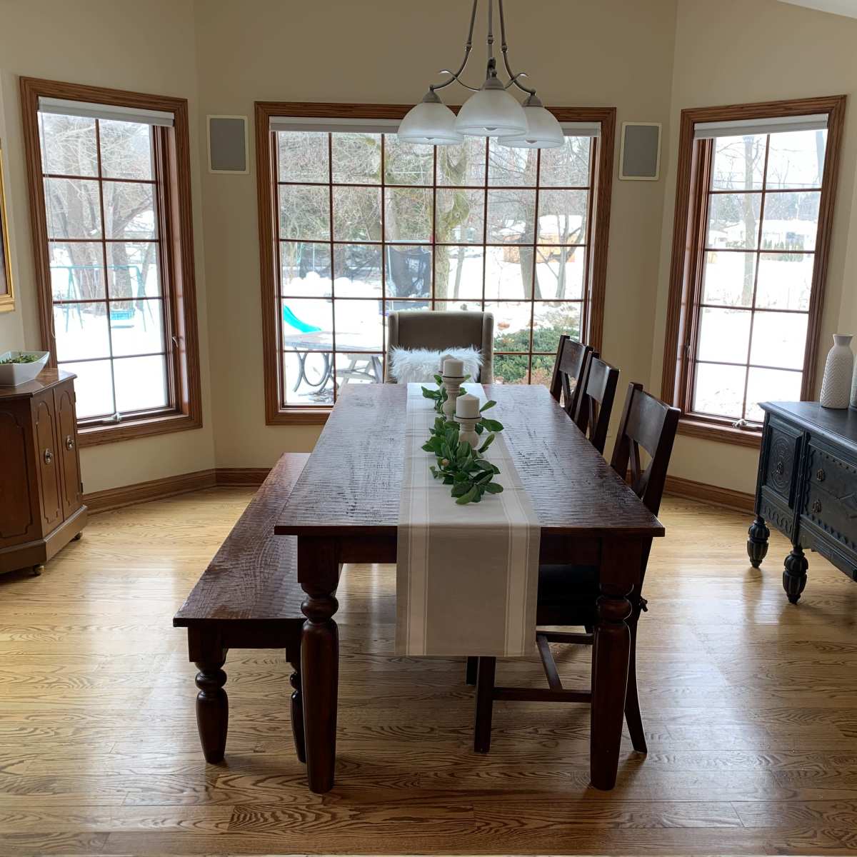 A wooden dining table with a runner and greenery sits in a well-lit room featuring large windows and a view of an outdoor space.