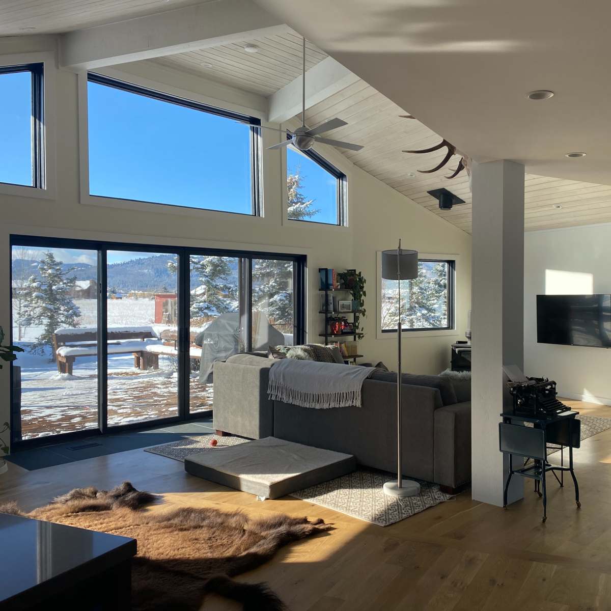 A modern living room with large windows overlooking a snowy landscape, furnished with a gray sofa and a television mounted on the wall.