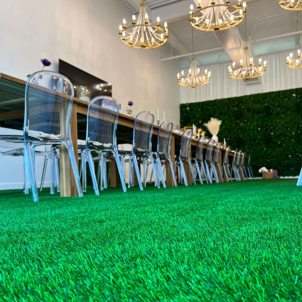 A long dining table set with clear chairs on a green artificial grass floor, illuminated by chandelier lights.