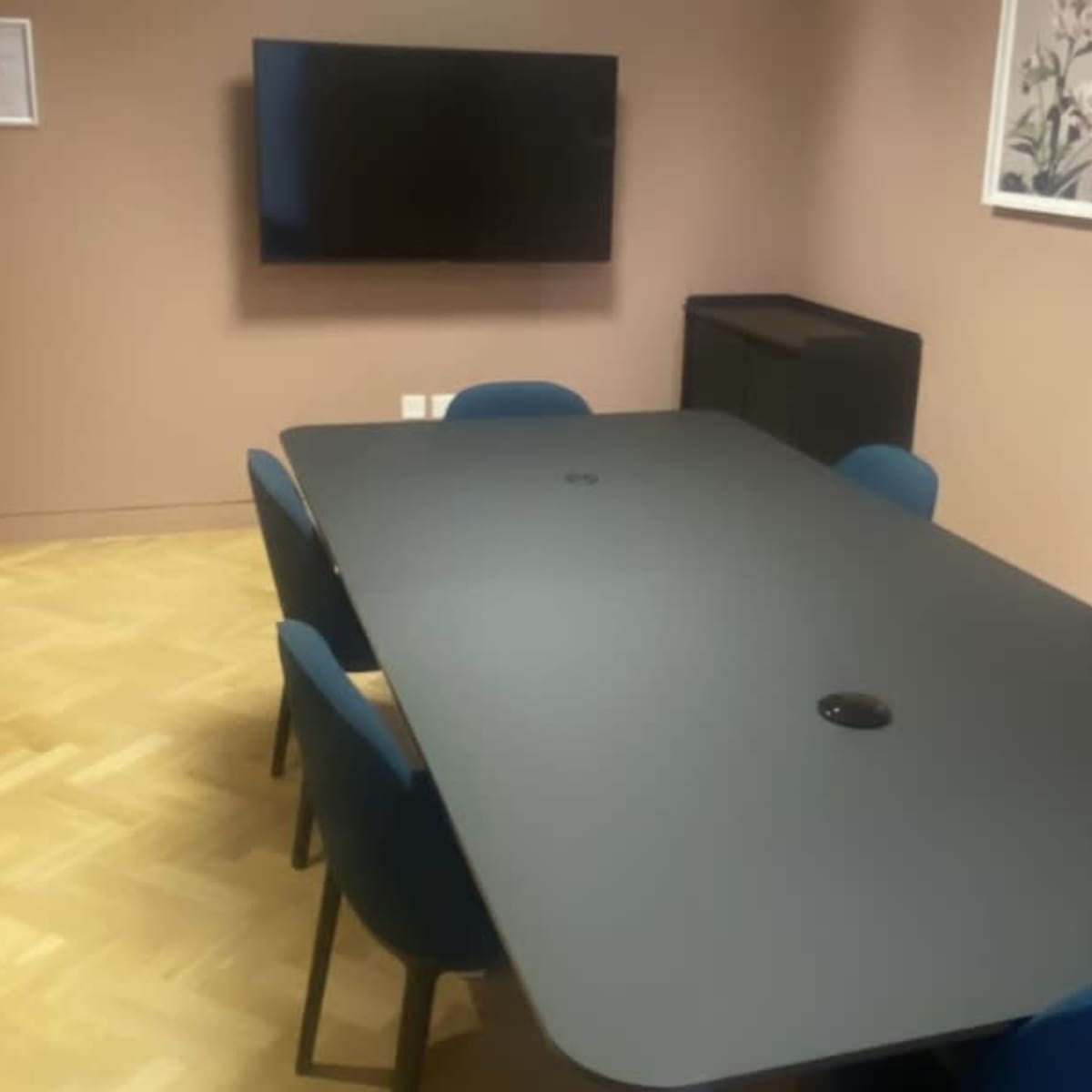 A large conference table with blue chairs is set up in a room featuring a television and framed wall art.