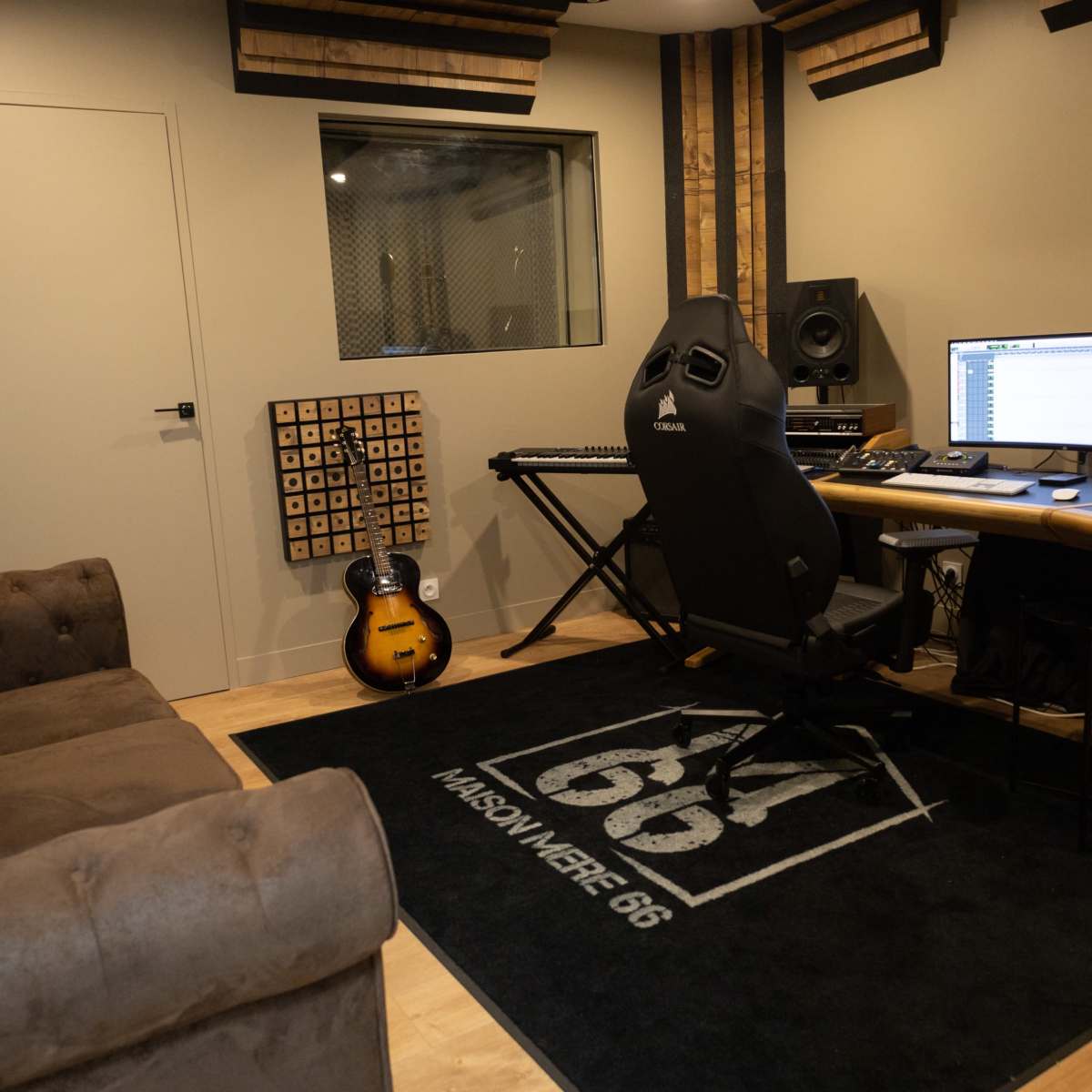 A music studio featuring a desk with a computer and audio equipment, a keyboard, an electric guitar against the wall, and a couch in front of a large rug.