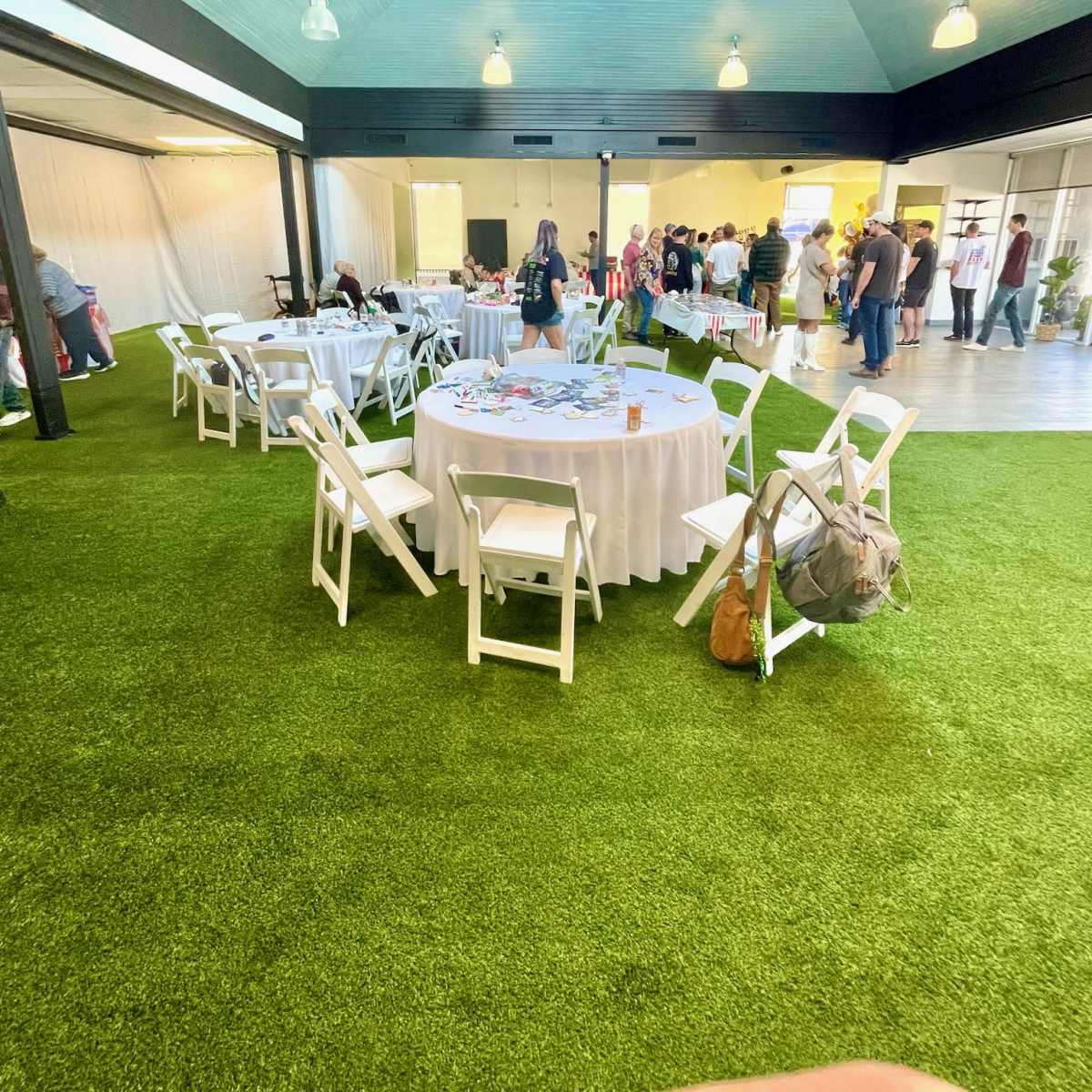 An event space with several round tables covered in white tablecloths, surrounded by white chairs, and a crowd of people engaging in activity in the background.