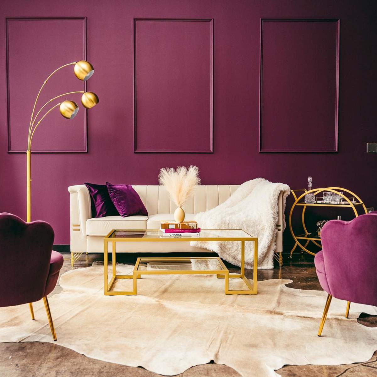 A modern living room with a purple wall, a white sofa, two violet chairs, and a gold coffee table on a light-colored rug.