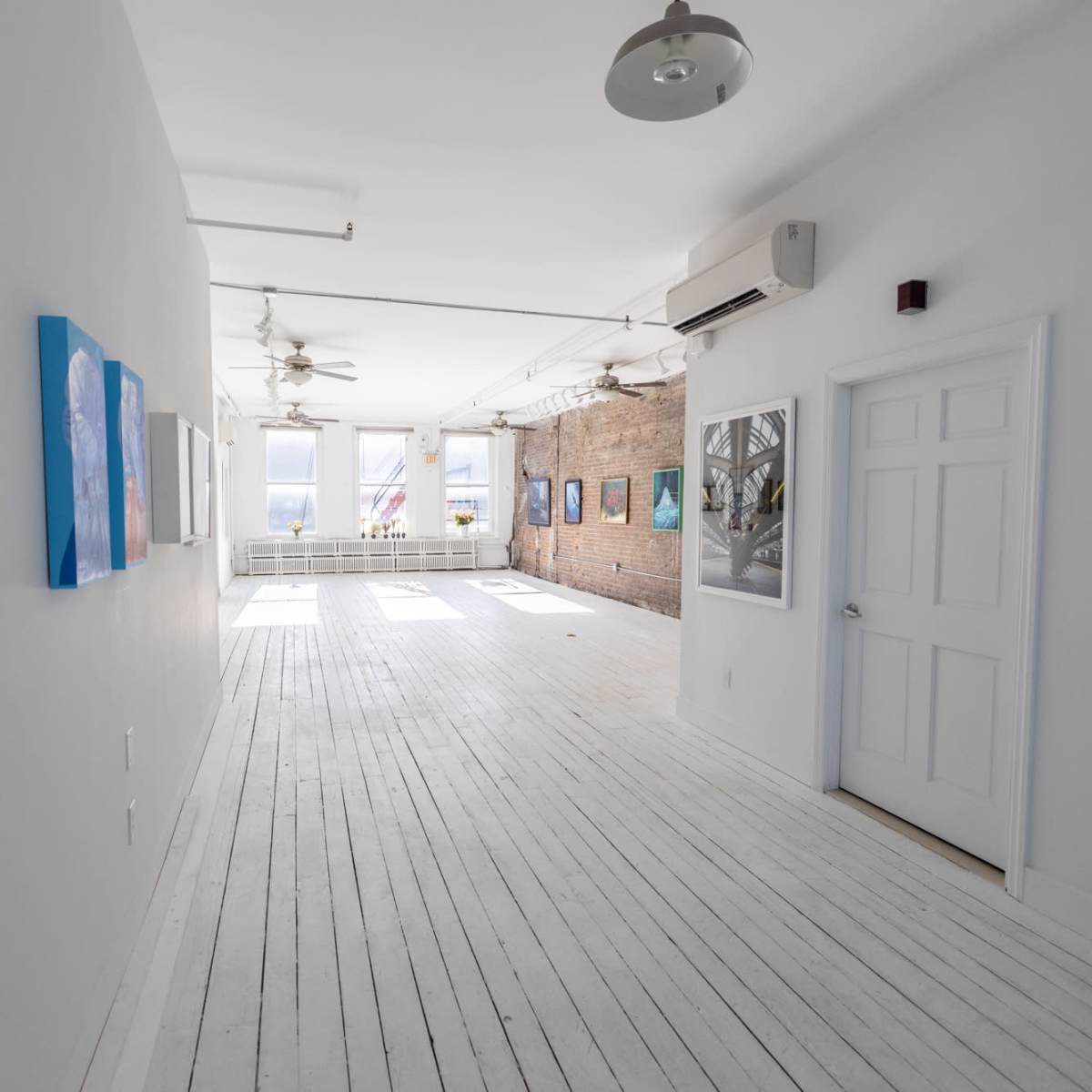 A bright, minimalist art gallery hallway featuring white wooden floors and several framed artworks on the walls.