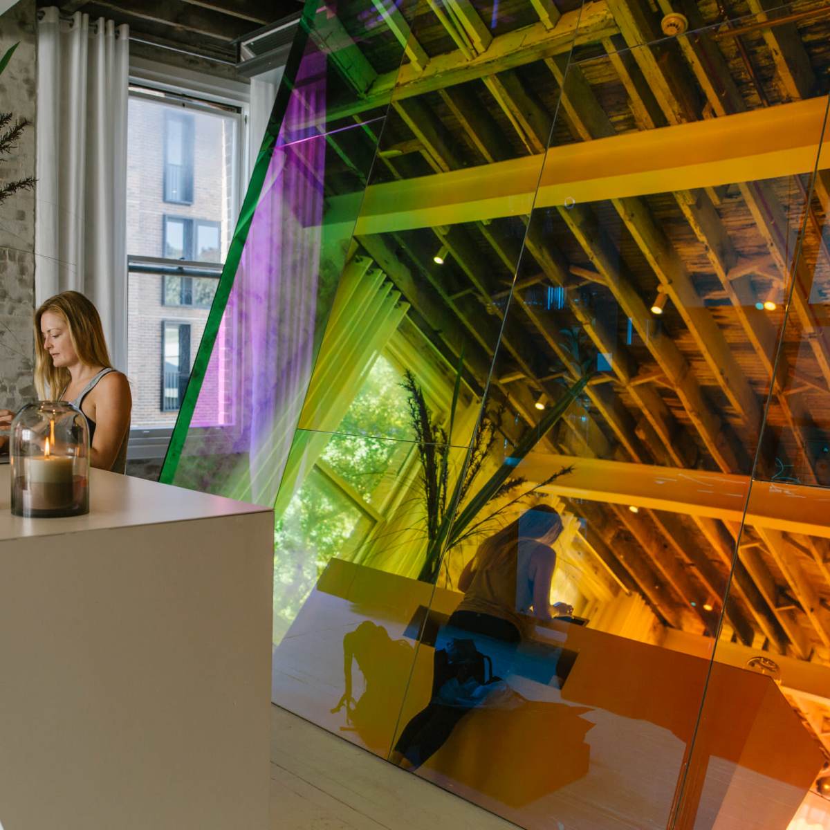 A woman is working on a laptop at a modern white desk in an office with a colorful reflective wall and large windows.