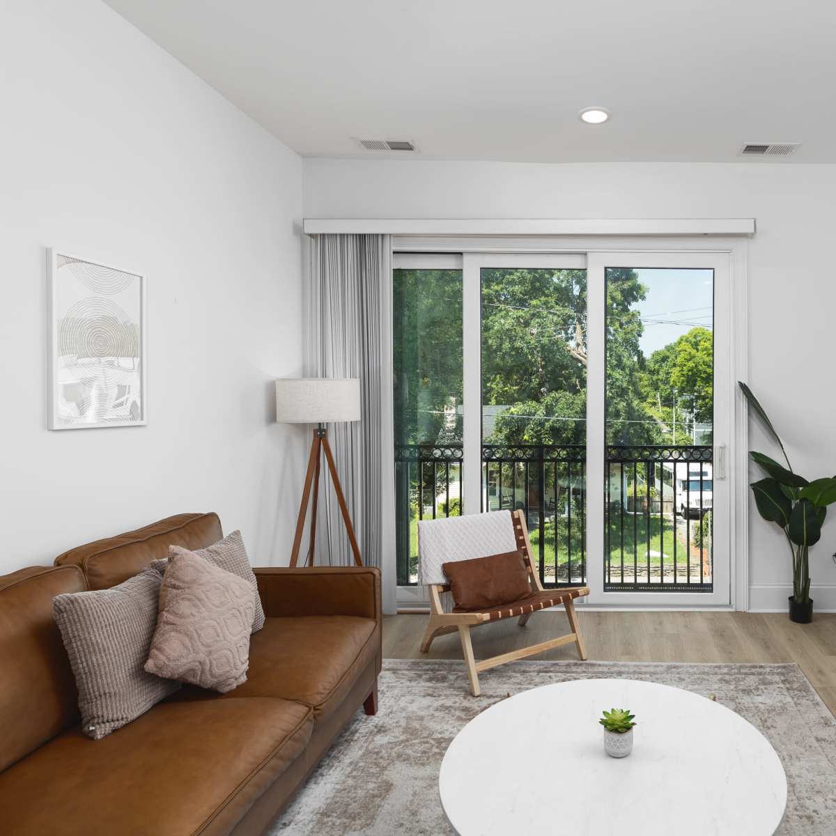 A modern living room features a brown leather sofa, a round coffee table, and a large window with a view of greenery outside.
