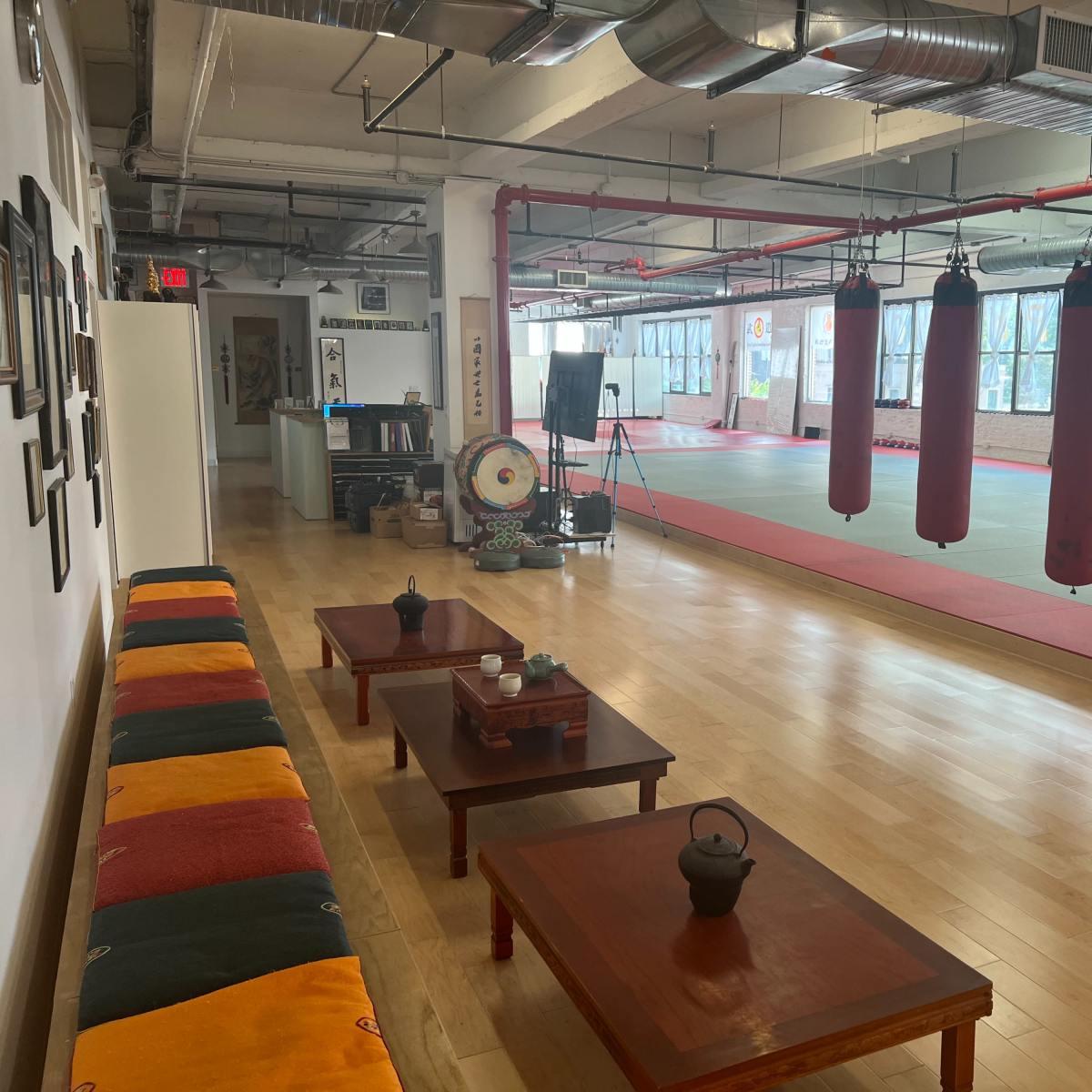 A spacious martial arts studio with hanging punching bags, wooden tables, and a seating area along the wall.