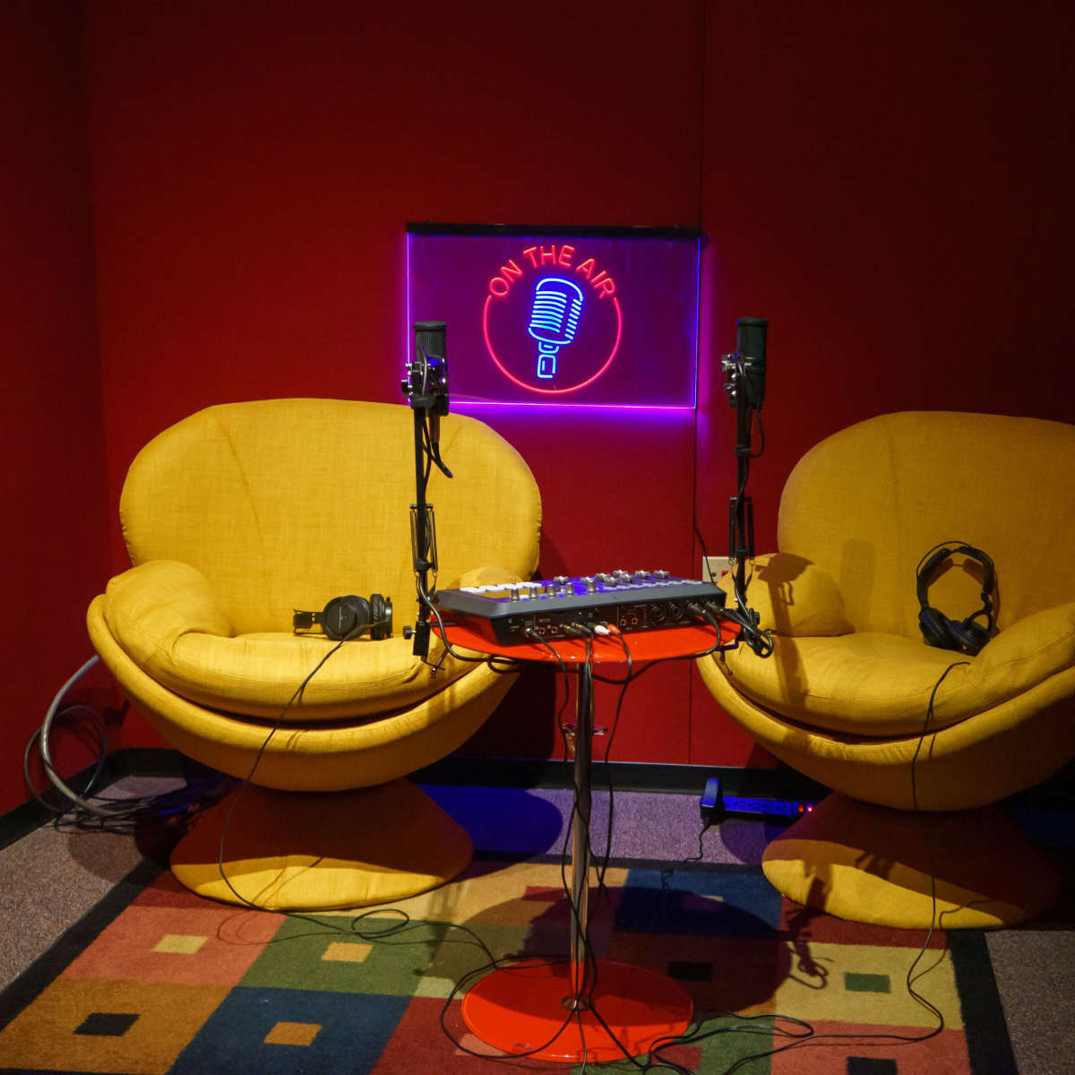 A small recording studio with two yellow chairs, a colorful carpet, a table with audio equipment, and a neon sign that says "ON THE AIR."