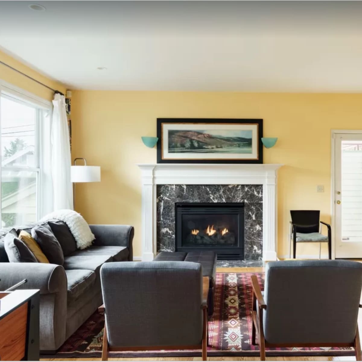 The room features a gray sofa, two armchairs, a fireplace with a marble surround, and a door leading outside, with natural light coming through large windows.