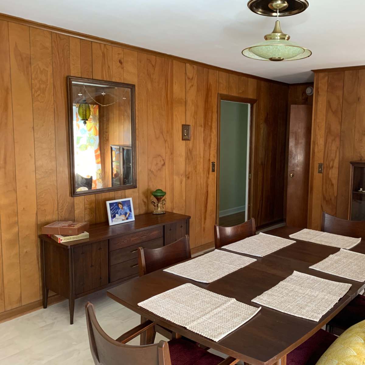 A dining room features a wooden table set with placemats, surrounded by chairs, with wood-paneled walls and a cabinet displaying various items.