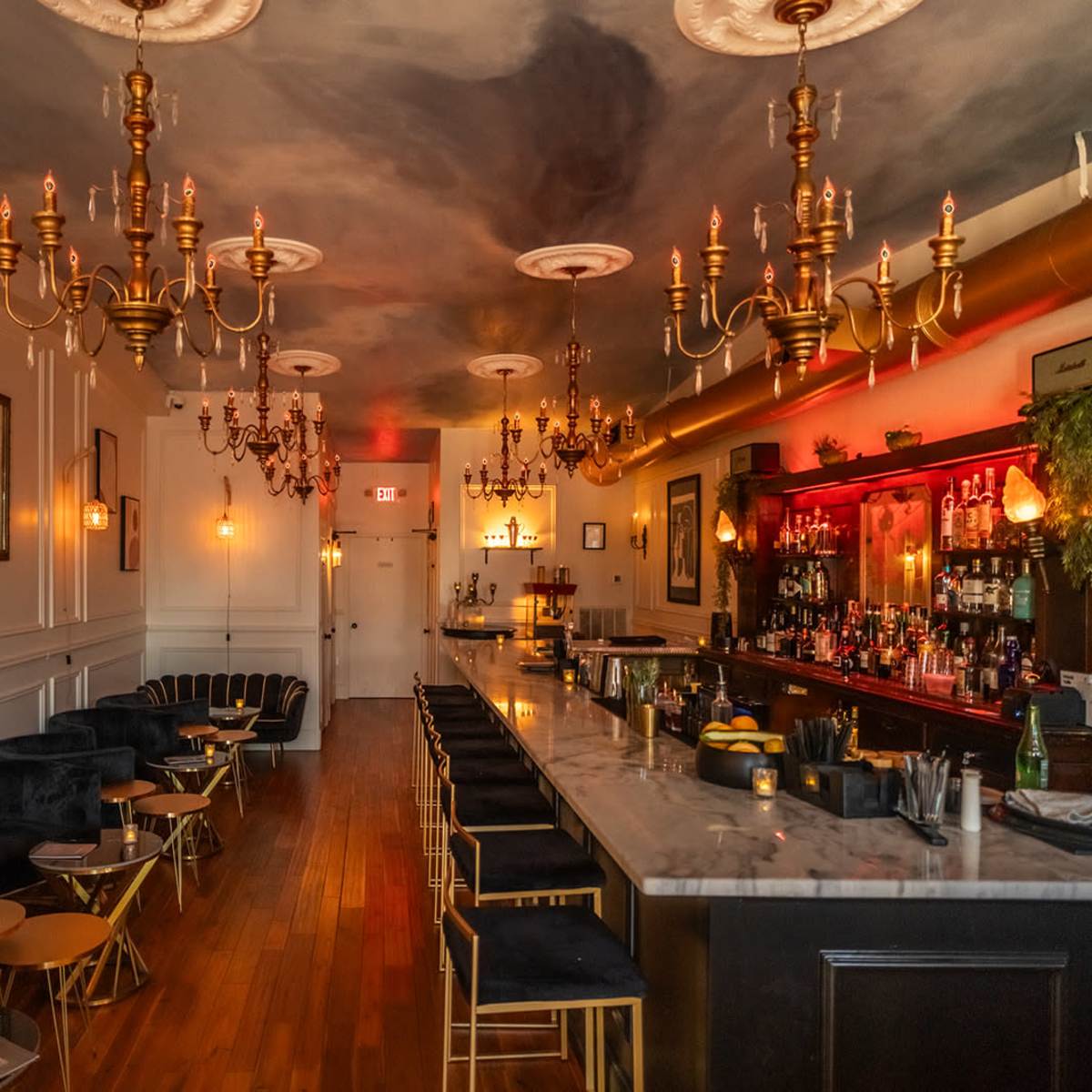 A stylish bar interior with a long marble counter, candle-lit tables, and ornate chandeliers hanging from the ceiling.