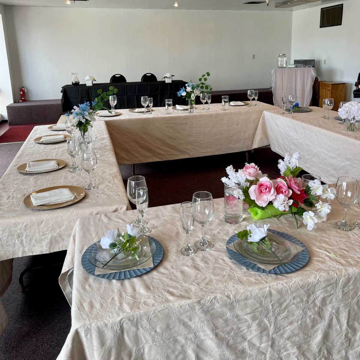 A spacious banquet room with multiple tables arranged in a square formation, each set with plates and glassware, and decorated with floral centerpieces.