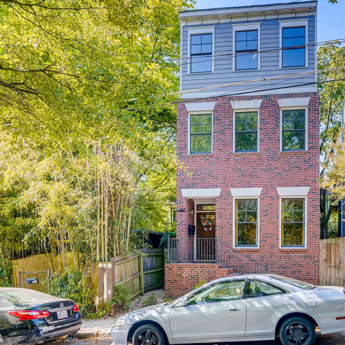 A three-story brick house with large windows is situated next to a green fence and various parked vehicles on a residential street.