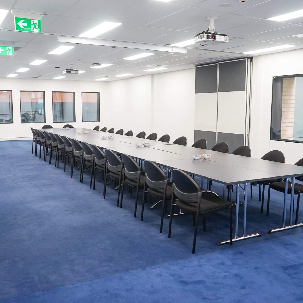 A long conference table with black chairs is arranged in a well-lit meeting room featuring windows and blue carpet.
