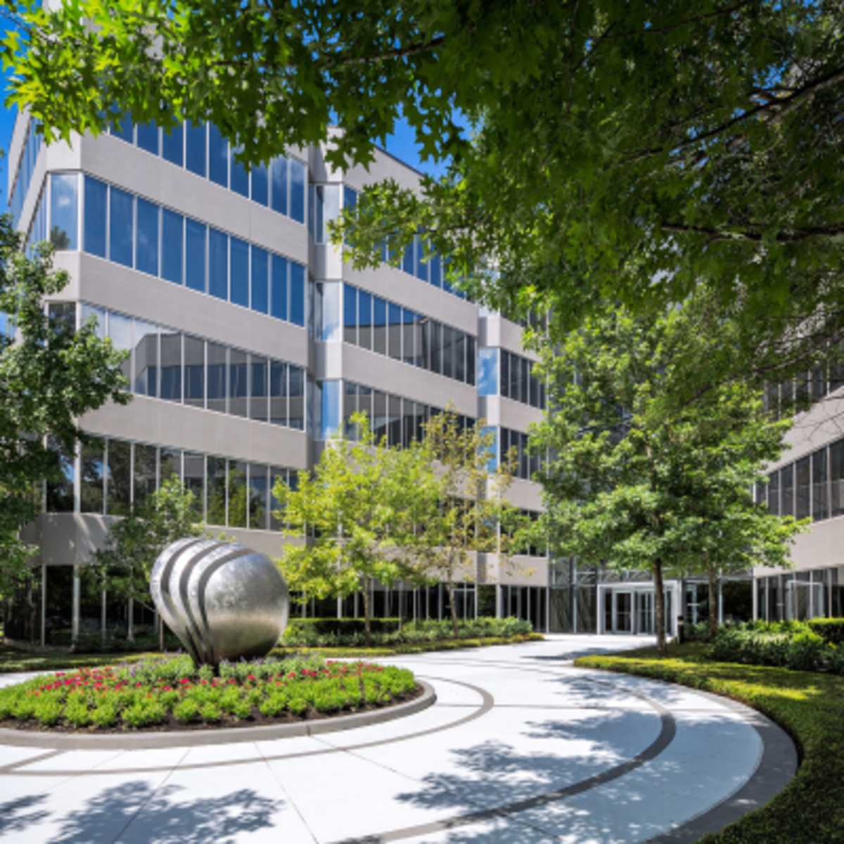 A modern office building is surrounded by greenery and has a decorative sculpture in a circular flowerbed at the entrance.