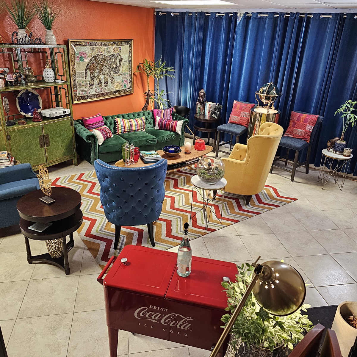 A brightly colored living room features a mix of furniture, including a green sofa, blue chairs, and a red Coca-Cola table, arranged around a patterned rug.
