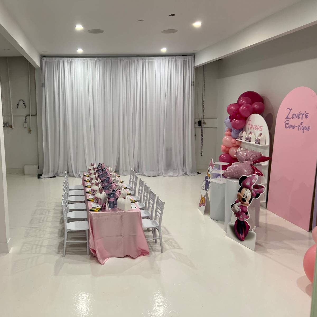 A decorated event space featuring a long dining table set with pink tableware and balloons, with white walls and a curtain backdrop.