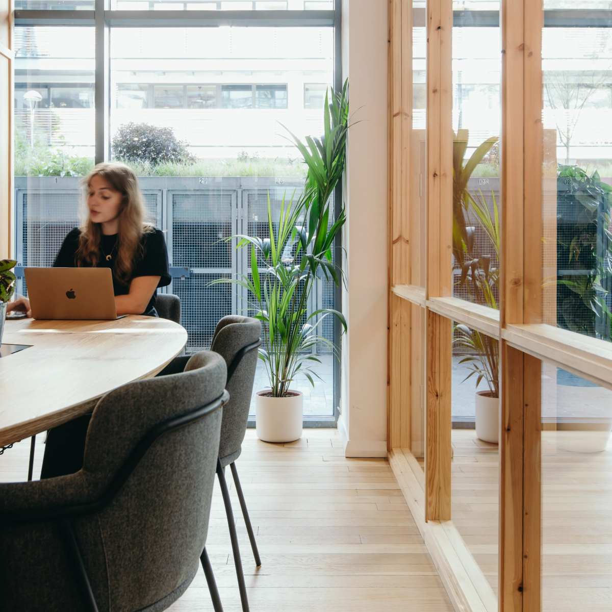 A woman works on a laptop at a wooden table in a modern office with large windows and potted plants.