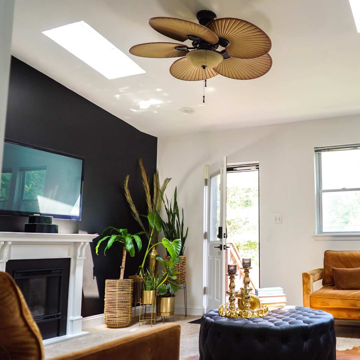 A modern living room features a brown leather chair, a black tufted ottoman, and large plants beside a white fireplace.