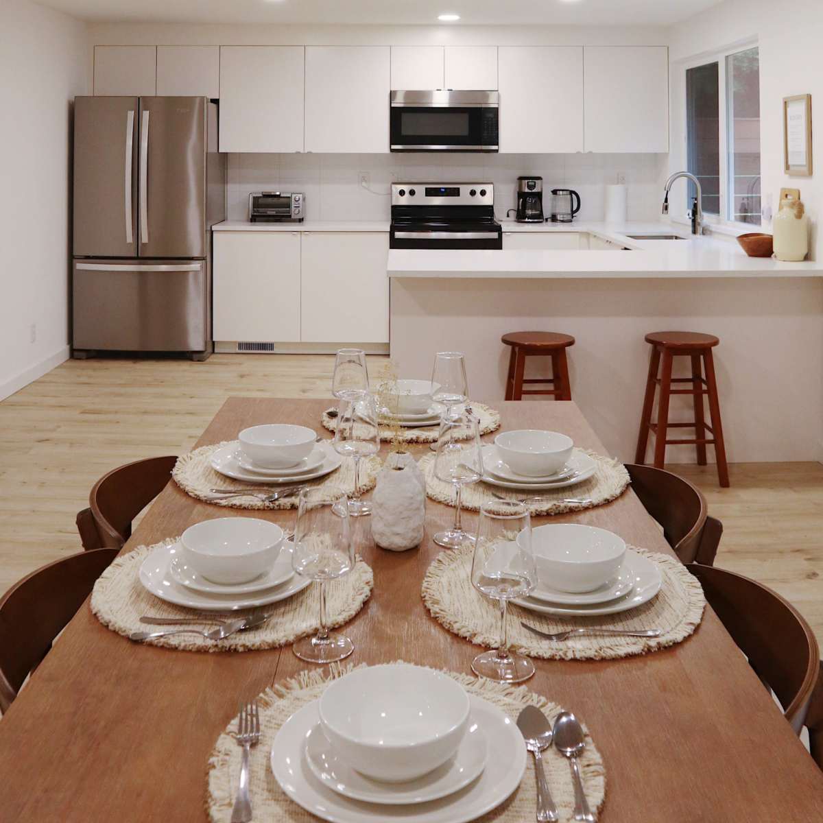 A modern kitchen features a dining table set with white plates and glassware, overlooking stainless steel appliances and a breakfast bar.