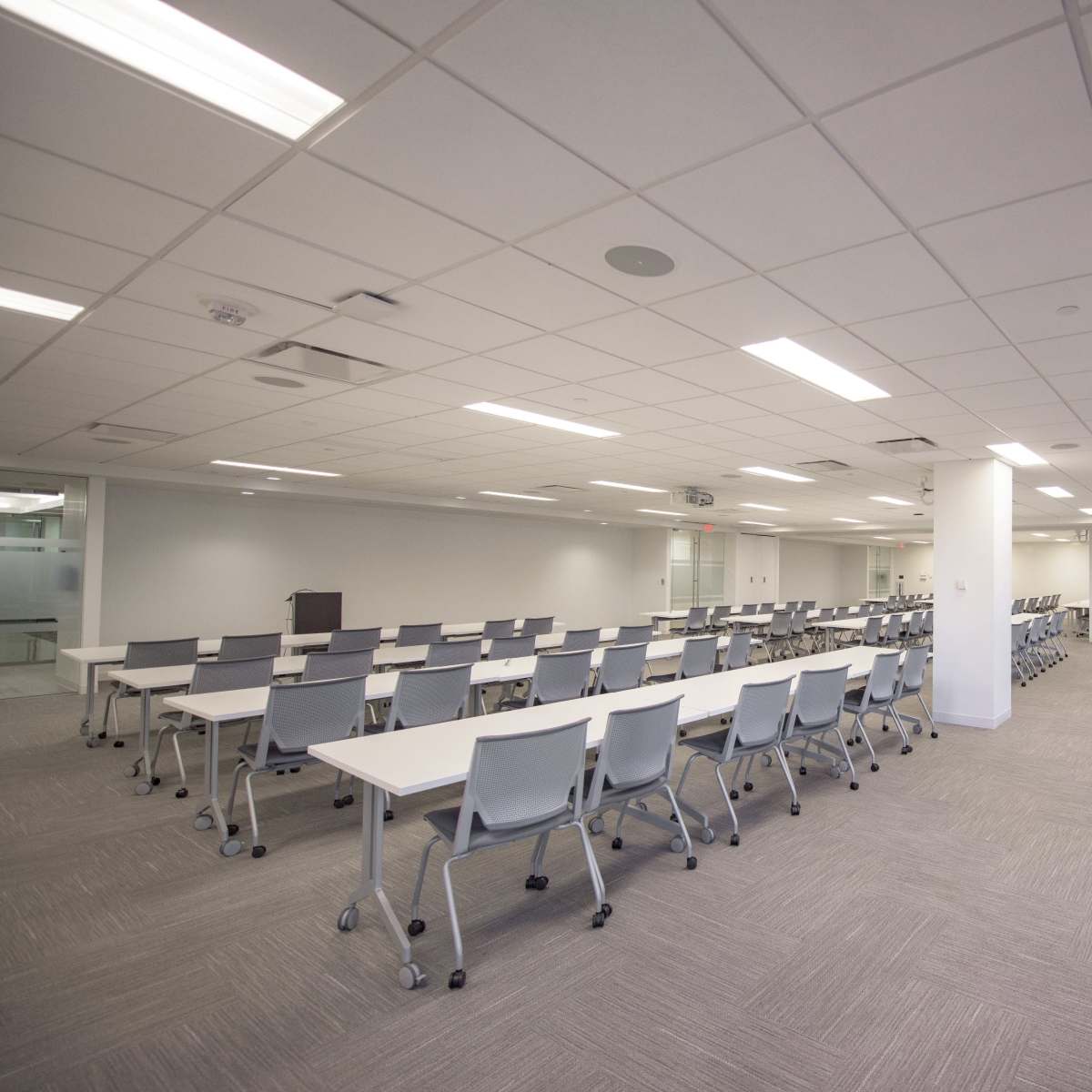 A spacious meeting room features rows of white tables and chairs, with large windows and neutral-colored carpeting.