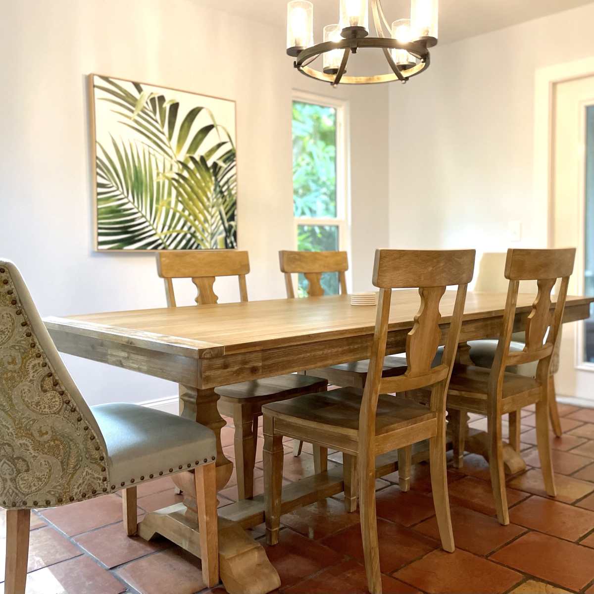 A wooden dining table is surrounded by six chairs in a well-lit room featuring large windows and a tropical-themed artwork on the wall.