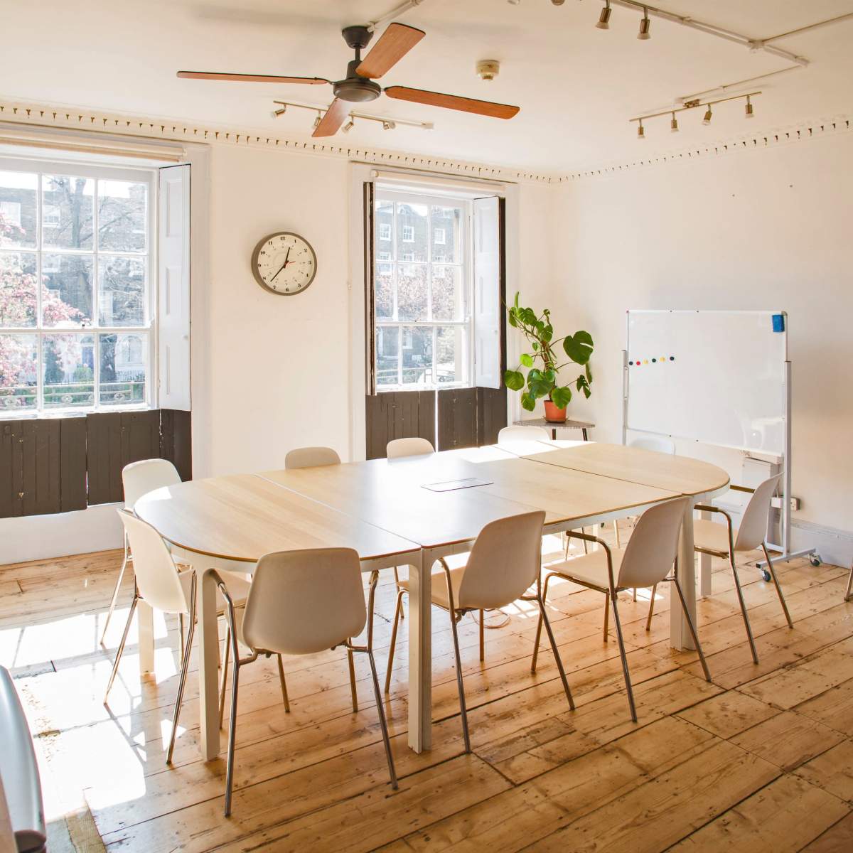 A bright meeting room with a large table surrounded by chairs, windows allowing natural light, and a clock on the wall.