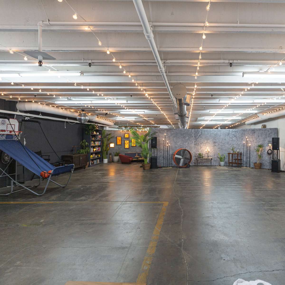 A spacious, industrial-style room with overhead lights, an open floor area, and various furnishings including a hammock, a television, and decorative plants.