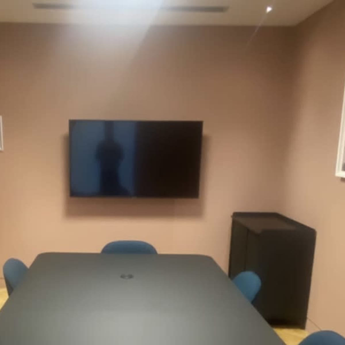 A conference room features a large table surrounded by blue chairs, a wall-mounted television, and a side cabinet.